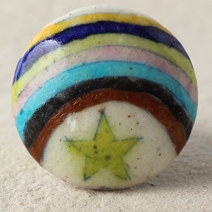 May include: A ceramic knob with a rainbow design and a yellow star in the center. The knob is round and has a white background.