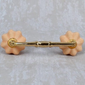 Może przedstawiać: Brzoskwiniowy uchwyt do szafki ze złotym drążkiem i kwiatowymi zakończeniami. Okucia przeznaczone są do mebli i charakteryzują się klasycznym, dekoracyjnym stylem. Uchwyt ma około 15 cm długości.