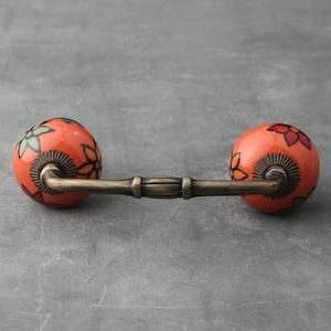 Può includere: Tiramaniglia per cassetto in ceramica arancione con un motivo floreale e un braccio in ottone. La maniglia è su uno sfondo grigio.