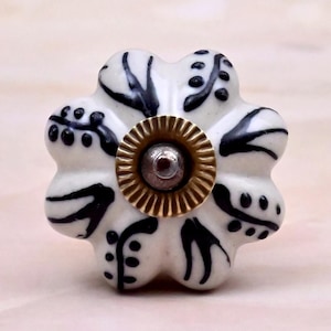 May include: A white ceramic cabinet knob shaped like a six-petaled flower with black hand-painted leaf and dot patterns. The centre of the knob features a circular brass-toned metal plate with a ridged edge and a small silver-toned screw head.