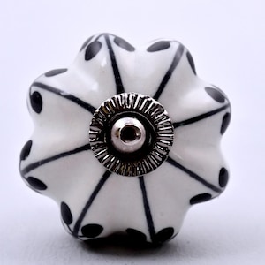 Op de afbeelding: Een witte keramische meubelknop met een geschulpte, bloemachtige vorm. De knop is voorzien van zwarte lijnen die vanuit het midden naar de randen uitstralen, met kleine zwarte bladachtige motieven aan de basis van elk bloemblad in het midden.