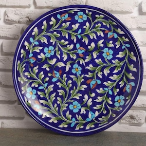 May include: A round ceramic plate featuring a deep blue background decorated with a repeating pattern of light blue flowers, yellow centers, and swirling green leaves. The design is symmetrical, with a dark blue border encircling the entire edge.