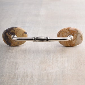 May include: A cabinet handle featuring a polished silver-tone metal bar connected to two oval-shaped, natural stone accents. The stone on the left displays dark brown and tan marbling, while the stone on the right features lighter tan and cream tones.