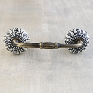 May include: A white ceramic drawer pull with a black floral design and a brass bar connecting the two knobs.