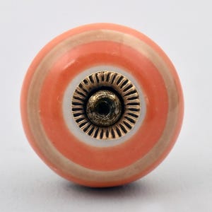 May include: A round cabinet knob featuring concentric circular bands of coral, beige, and white. The center of the knob is fitted with a circular, metallic brass-toned hardware piece that has a textured, ridged edge and a central screw hole.