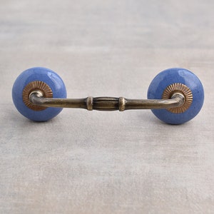 May include: A blue ceramic drawer pull with a brass bar connecting the two knobs. The knobs have a gold trim around the edge.