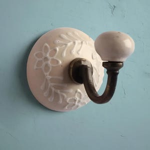 Puede incluir: Gancho de pared de cerámica blanca con un diseño floral y un gancho de metal. El gancho está unido a una base de cerámica blanca con un diseño floral.