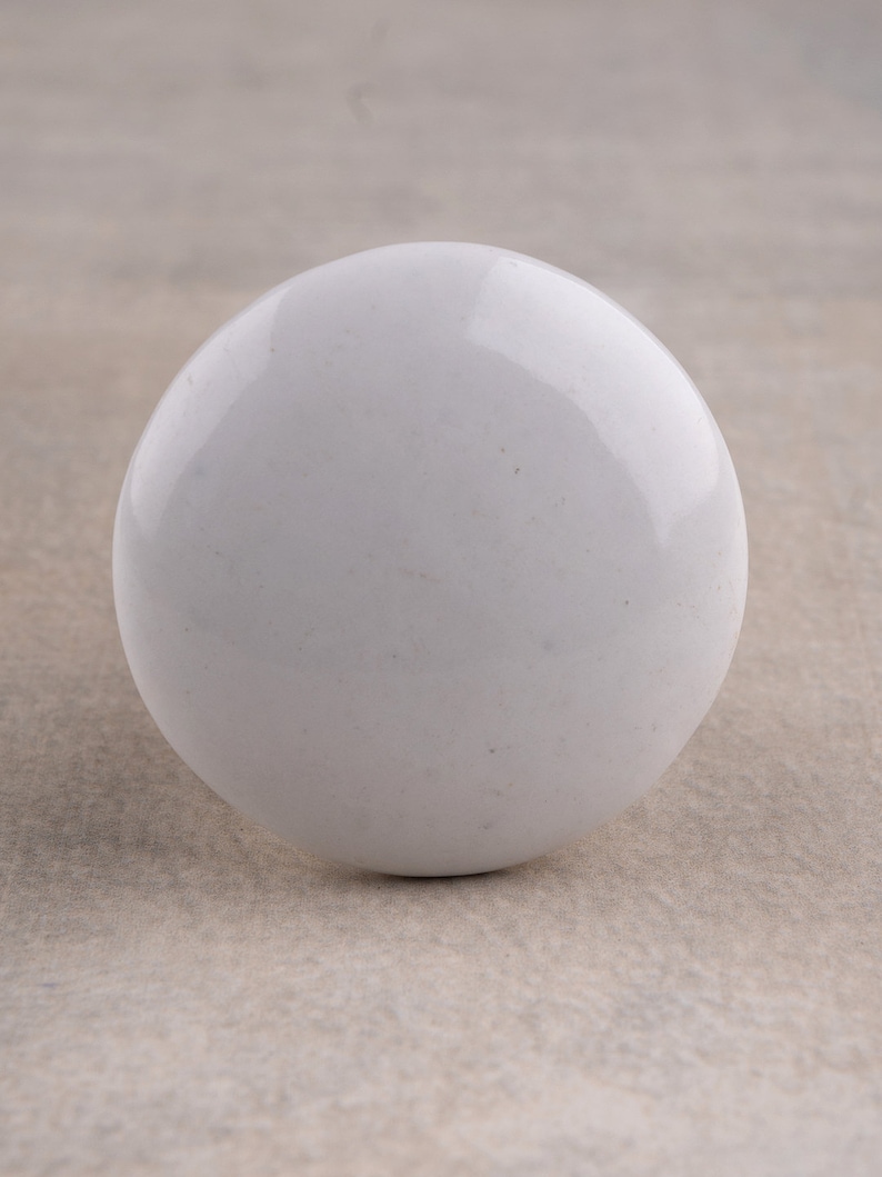 May include: A smooth, spherical white knob with a glossy, reflective finish. The object has a uniform, solid white colour throughout its rounded surface, presenting a clean and simple circular shape that is consistent from this front-facing perspective.