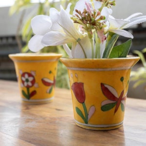 May include: Two yellow ceramic flower pots with colorful floral and butterfly designs. The pots are on a wooden surface and one pot has white flowers inside.