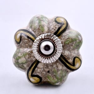 Puede incluir: Un tirador de mueble de cerámica con forma de flor y acabado moteado en gris y verde. La superficie presenta patrones de líneas negras y amarillas en espiral. Una base metálica plateada con borde estriado está centrada en la parte frontal.