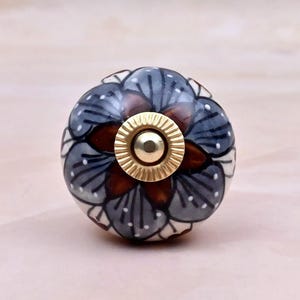 Op de afbeelding: Een ronde keramische meubelknop met een handgeschilderd bloemmotief in leigrijs, donkerbruin en wit. Het midden van de knop is voorzien van een cirkelvormig, geribbeld beslag van goudkleurig metaal met een gladde, gepolijste afwerking.