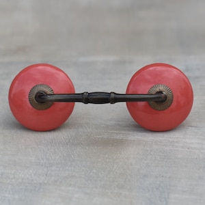 May include: A pair of coral-colored ceramic cabinet knobs with a dark bronze-colored connecting bar. The knobs are round with a slightly textured surface and are designed for easy grip and installation. The hardware is suitable for drawers and cabinets.