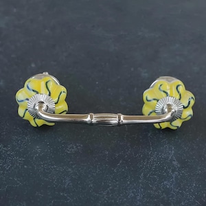 May include: A yellow and blue ceramic drawer pull with a silver metal arm. The pull has a floral design.