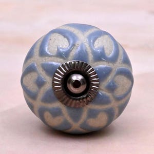 May include: A round ceramic cabinet knob featuring a light blue and off-white repeating heart-shaped pattern. The centre of the knob has a circular silver-toned metal hardware piece with a ridged edge and a small, smooth, raised metal dome.