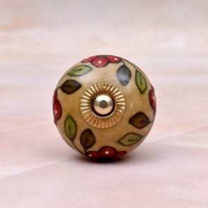May include: A round ceramic cabinet knob with a cream-colored base featuring a hand-painted floral pattern of red flowers and green leaves. The center of the knob has a circular gold-toned metal hardware piece with a ridged edge and a smooth center.