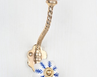 May include: A wall-mounted hook featuring a gold-toned twisted metal arm and two white ceramic knobs. Each knob is shaped like a flower and decorated with blue painted leaf patterns and a small gold-toned center cap for mounting items.