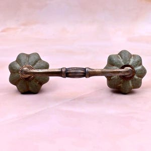 May include: A drawer pull featuring a horizontal metallic handle with a dark, weathered finish, connected to two round, fluted ceramic knobs in a muted sage green colour. The handle has a central decorative band and ribbed circular bases.