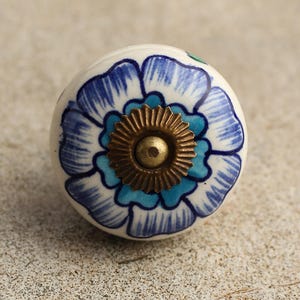 Könnte beinhalten: Ein weißer Keramik-Knopf mit einem blauen und türkisfarbenen Blumendesign. Der Knopf hat eine Messingmitte mit einem dekorativen Ring.