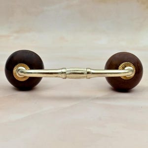 May include: A drawer pull featuring a horizontal gold-toned metal bar with a central decorative segment, connected to two dark brown spherical wooden knobs on each end, each accented with a small gold-toned metal circular plate at the base.