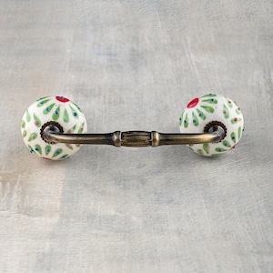 May include: A white ceramic drawer pull with a green and red floral design. The pull has a brass bar connecting the two knobs.
