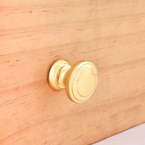 May include: A gold-toned cabinet knob with a round, slightly raised design. The knob is mounted on a light brown wooden surface.