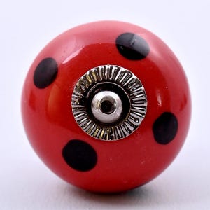 May include: A round, red ceramic cabinet knob featuring black polka dots. The center of the knob has a circular, silver-colored metal hardware piece with a ridged, sunburst-like texture surrounding a small, central mounting hole for installation.