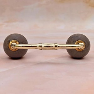 Pode incluir: Um puxador de móvel com dois botões redondos de pedra cinza mate ligados por uma barra de metal dourado polido. A barra tem um detalhe central canelado e é fixada a cada botão com uma pequena placa metálica circular dourada com estrias.