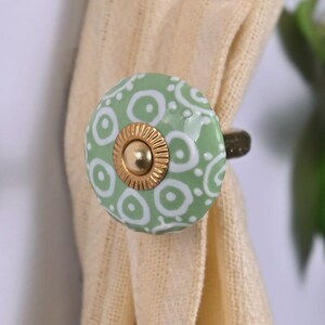 May include: A round, light green ceramic curtain tieback featuring a white circular pattern and a central gold-colored metal disc with a ridged edge. The tieback is shown holding a piece of light beige fabric in place, securing it with its hardware.