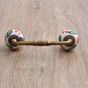 May include: A decorative cabinet pull with a brass handle and two white ceramic knobs. The knobs are adorned with a floral pattern in orange, green, and blue. The pull is designed for furniture and adds a touch of elegance.