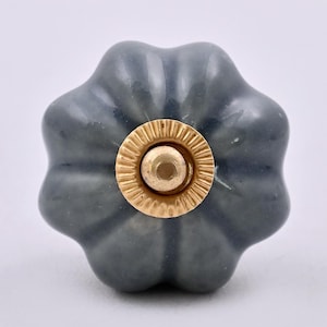 Puede incluir: Un tirador de mueble con forma de calabaza, acabado en gris oscuro brillante y una placa circular central de metal dorado con borde estriado. El tirador presenta una superficie lisa y redondeada con marcadas hendiduras verticales.