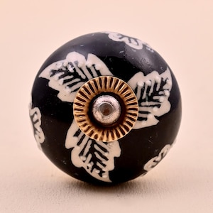 Puede incluir: Un pomo de mueble redondo de cerámica negra con un patrón de hojas blancas en su superficie. El centro del pomo está acentuado con una placa metálica circular de tono dorado y una pequeña cabeza de tornillo plateada, contrastando con el acabado.