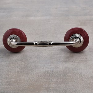 May include: A cabinet pull with a polished chrome finish and two round, dark red ceramic knobs. The knobs have a decorative silver trim. The handle is a simple, elegant design.