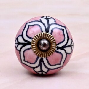 May include: A round ceramic cabinet knob featuring a soft pink base color with a hand-painted white and black floral pattern. The center of the knob is accented with a circular, ridged brass-toned metal fitting and a small central screw head.