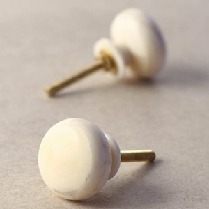 May include: Two white ceramic cabinet knobs with gold metal screws. The knobs are round and have a slightly textured surface.