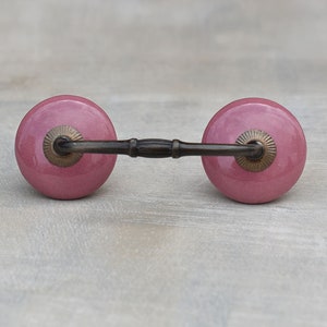 Pode incluir: Um par de puxadores de gaveta de cerâmica rosa com uma barra de metal que os conecta. Os puxadores têm um design circular ligeiramente elevado.