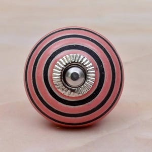May include: A round ceramic cabinet knob featuring a series of concentric circles in alternating shades of salmon pink and black. The centre of the knob is adorned with a metallic silver-toned circular plate with a fluted edge and a central bolt.