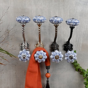 Op de afbeelding: Vier decoratieve wandhaken met blauwe en witte keramische knoppen. De haken zijn gemaakt van metaal en hebben een gedraaid ontwerp. Een rode sjaal hangt aan een van de haken.