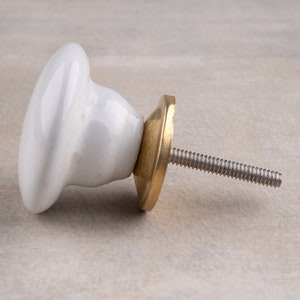 May include: A white ceramic cabinet knob with a smooth, rounded top and a tapered base. The knob features a circular brass-toned metal backplate and a threaded metal bolt extending from the centre, designed for attachment to furniture or doors.