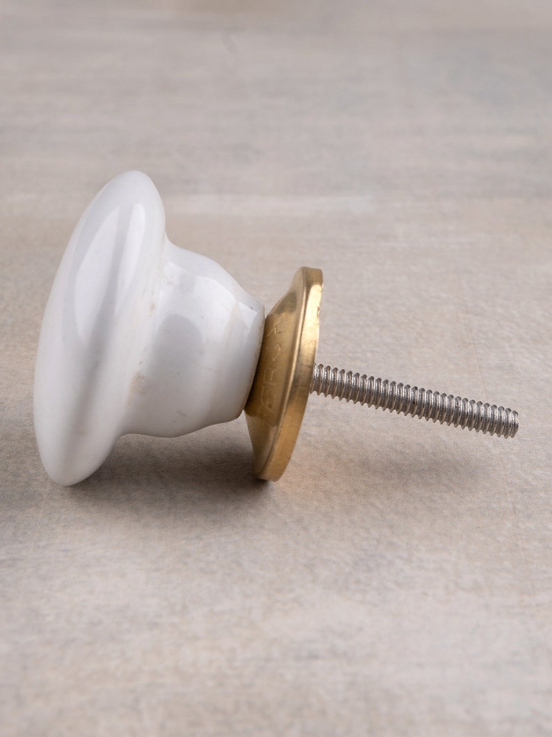 May include: A white ceramic cabinet knob with a smooth, rounded top and a tapered base. The knob features a circular brass-toned metal backplate and a threaded metal bolt extending from the centre, designed for attachment to furniture or doors.