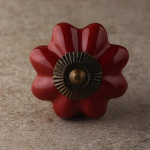 May include: A red ceramic cabinet knob with a flower-like design. The knob features a dark bronze-colored metal center with a brass-colored center. The knob is a decorative accent for furniture.