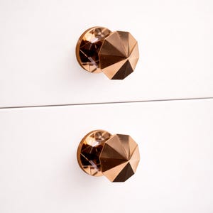 May include: Two rose gold cabinet knobs with a faceted, geometric design. The knobs are mounted on a white surface, and the design creates a play of light and shadow. These decorative knobs add a touch of elegance.