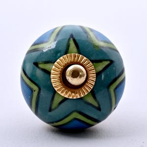 Puede incluir: Un pomo de mueble redondo de cerámica con un patrón de estrella azul y verde con contornos negros. El centro del pomo está adornado con una pieza de metal dorado circular que presenta un borde estriado y festoneado y una parte superior redondeada.