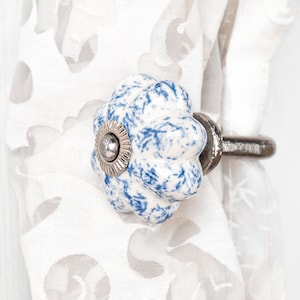 Może przedstawiać: Ceramiczny gałka w kolorze niebieskim i białym z srebrnym środkiem, przymocowana do srebrnego metalowego haka. Hak jest przymocowany do białej zasłony z kwiatowym wzorem.