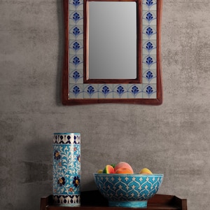 Könnte beinhalten: Eine blaue und weiße Keramikvase mit Blumendesigns steht auf einem Holztisch mit zwei Schubladen. Eine blaue und weiße Keramikschale mit Obst steht auf dem Tisch neben der Vase. Ein Spiegel mit einem blauen und weißen Fliesenrahmen hängt an der Wand hinter dem Tisch.