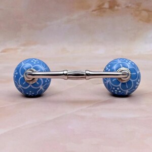 Può includere: Una maniglia per mobili con due pomelli sferici in ceramica blu con motivo floreale bianco, collegati da una barra in metallo color argento lucido. La ferramenta include basi circolari argentate nei punti di giunzione della barra.