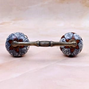 Peut inclure: Une poignée de tiroir composée de deux boutons en céramique ronds reliés par une barre horizontale en laiton antique. Chaque bouton présente un motif floral avec des pétales rouge foncé, des accents gris clair et des points blancs.