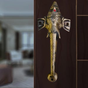 May include: A gold-toned door handle shaped like the head of the Hindu deity Ganesha. The handle has a red jewel in the center of the forehead and a detailed design on the head and trunk.