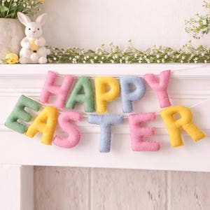 May include: A pastel-coloured felt banner spelling "HAPPY EASTER". The letters are strung on a white string. A ceramic bunny and floral arrangement are in the background. The banner is displayed on a white surface.