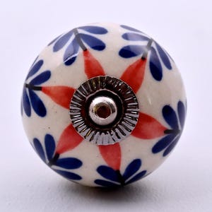 May include: A round ceramic cabinet knob with a cream-coloured base featuring a repeating pattern of blue petal-like shapes and orange star-shaped accents. The centre of the knob has a circular silver-toned metal hardware piece with ridges.