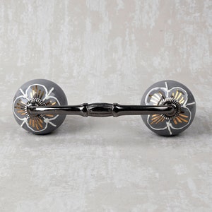 Puede incluir: Un tirador decorativo para armario con acabado gris oscuro. El tirador presenta dos pomos redondos con un diseño floral en blanco y dorado. La barra de conexión es de color metálico oscuro. Para muebles y decoración del hogar.
